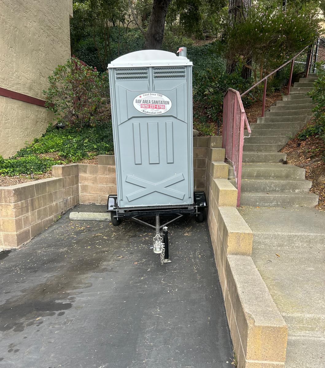 Trailer-mounted Bay Area Sanitation porta potty on a driveway beside outdoor stairs, available in Crockett