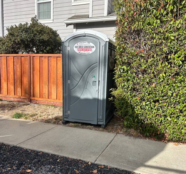 Porta potty between a wood fence and hedge outside a home, available in Discovery Bay